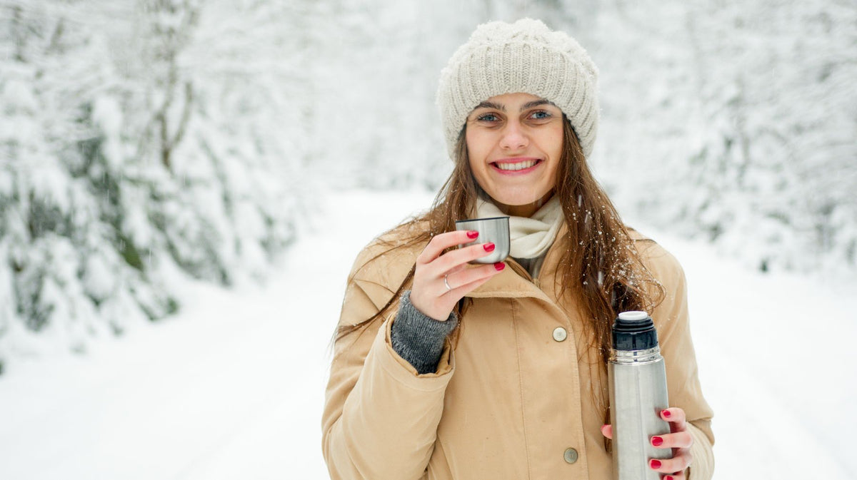 Smiling woman in a winter coat and knit hat holding a hot drink in the snow